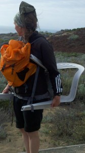 A woman with an orange backpack carries a plastic chair and looks out toward the sand dunes.