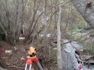 Staff uses an autolevel to determine the elevation of a series of benchmarks. An autolevel a piece of surveying equipment that is essentially a telescope. You can see a staff member holding a stadia rod (giant rule) in the trees.