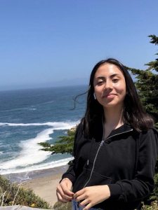 Anahi Peralta Cordova stands inn front of a tree by the ocean. She wears a black zip-up sweatshirt. She is looking at the camera. 