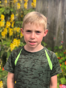 Seamus Wolf looks at the camera. he has brown hair, wears a grey shirt with green diagonal stripes at the shoulders, and stands in front of yellow flowers, green foliage, and a brown fence.