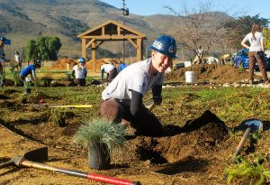 Corpsmember posing in a hard hat while planting drought-resistant plants in SLO County.