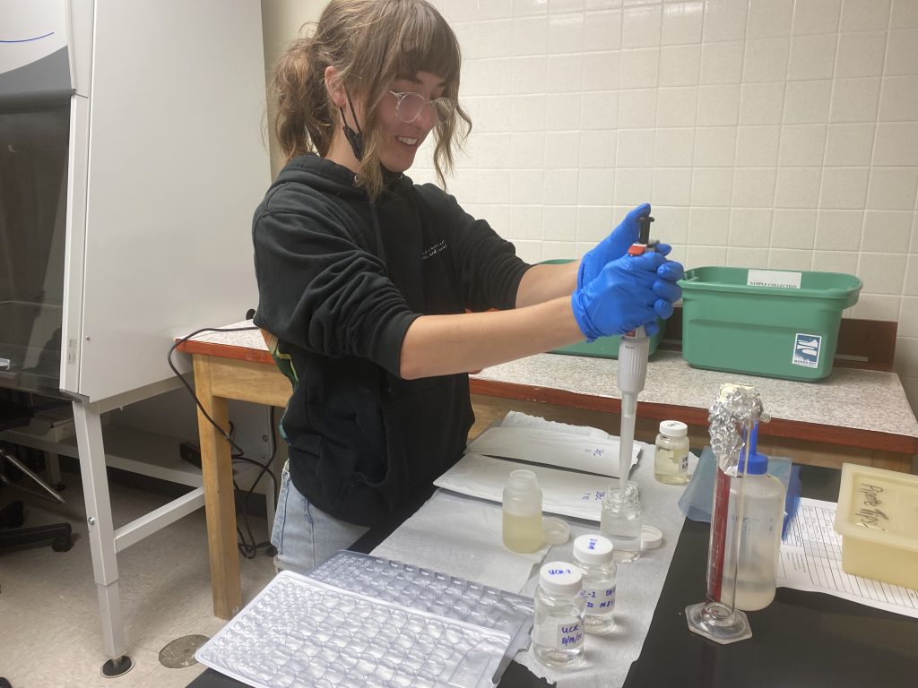 A young person with brown hair and clear glasses uses a pipette in a lab setting.
