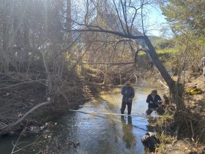 Two people wearing waders are standing knee-deep in a creek. One appears to be taking measurement, and the other is writing on a clipboard.