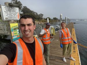 Three people wearing orange safety vests stand on a dock.