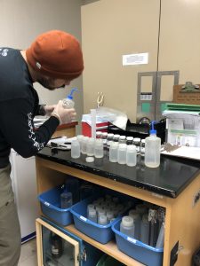 A person with an orange beanie stands in front of a row of plastic bottles in a lab setting. They have a squeeze bottle in their hands.