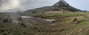 A panoramic image of a floodplain. There is a noticeable lack of plants, and evidence of erosion.